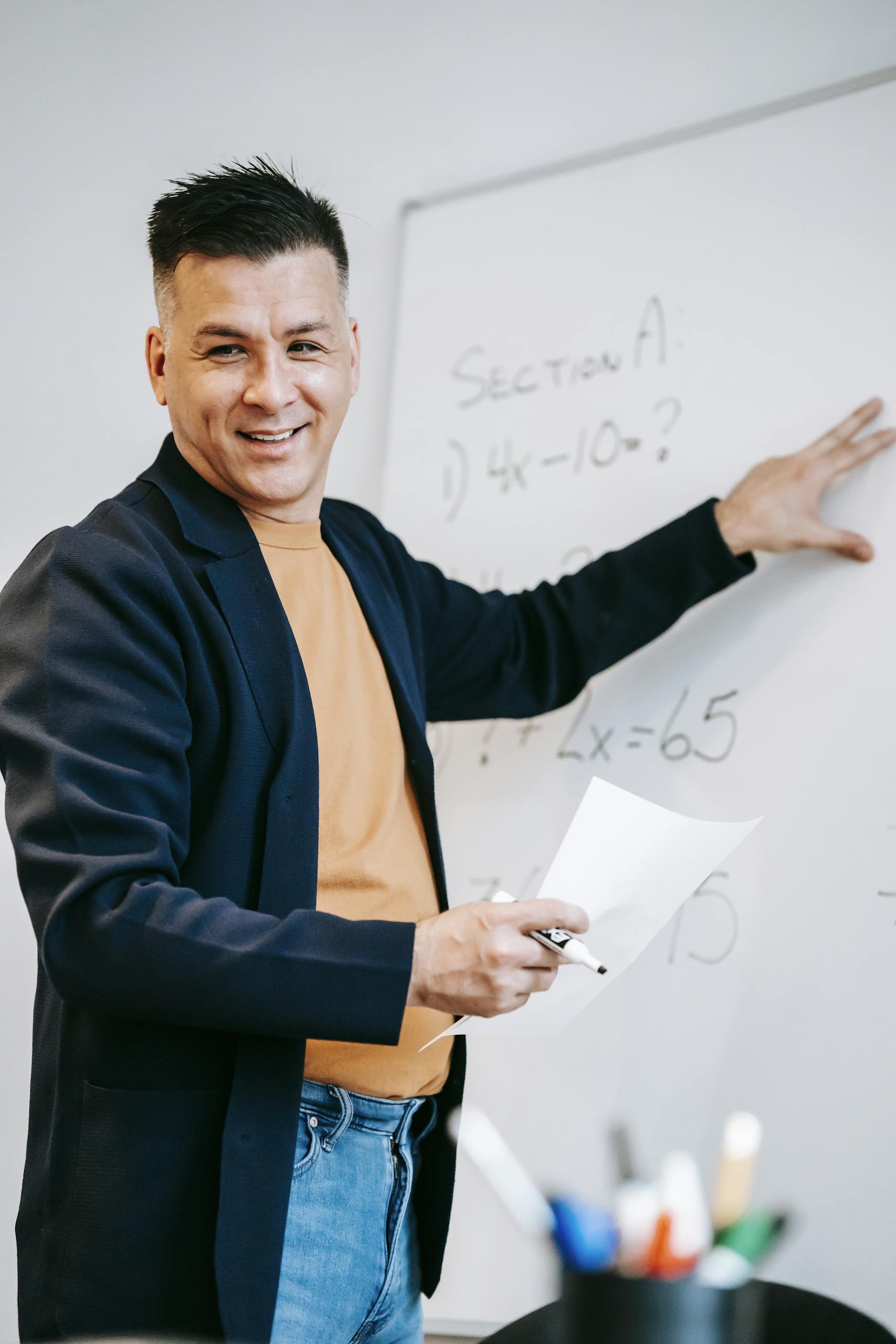 Friendly teacher explains equations on a whiteboard in a classroom setting.
